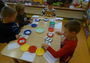 Czwórka dzieci siedzi przy stole, przed każdym dzieckiem leżą po trzy plastikowe talerzyki. Dzieci trzymają w ręku obrazki ptaków, segregują je na odlatujące, przylatujące i pozostające w Polsce.